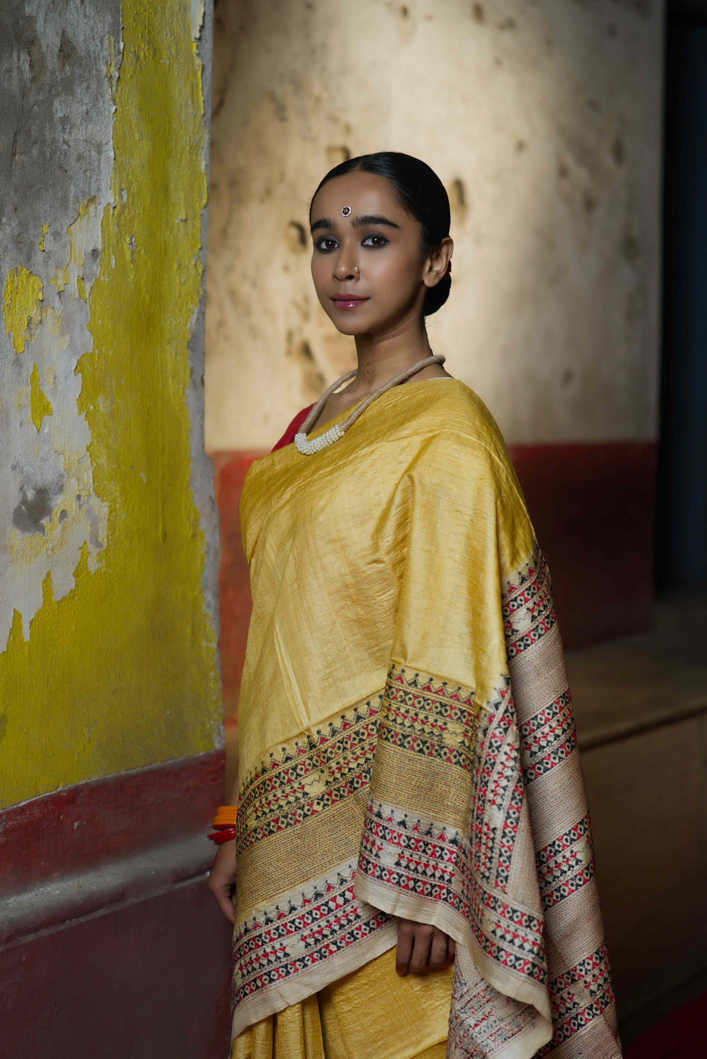 A woman wearing a handwoven yellow tussar saree with kantha work standing against a wall with colorful graffiti.