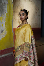 A woman wearing a handwoven yellow tussar saree with kantha work standing against a wall with colorful graffiti.